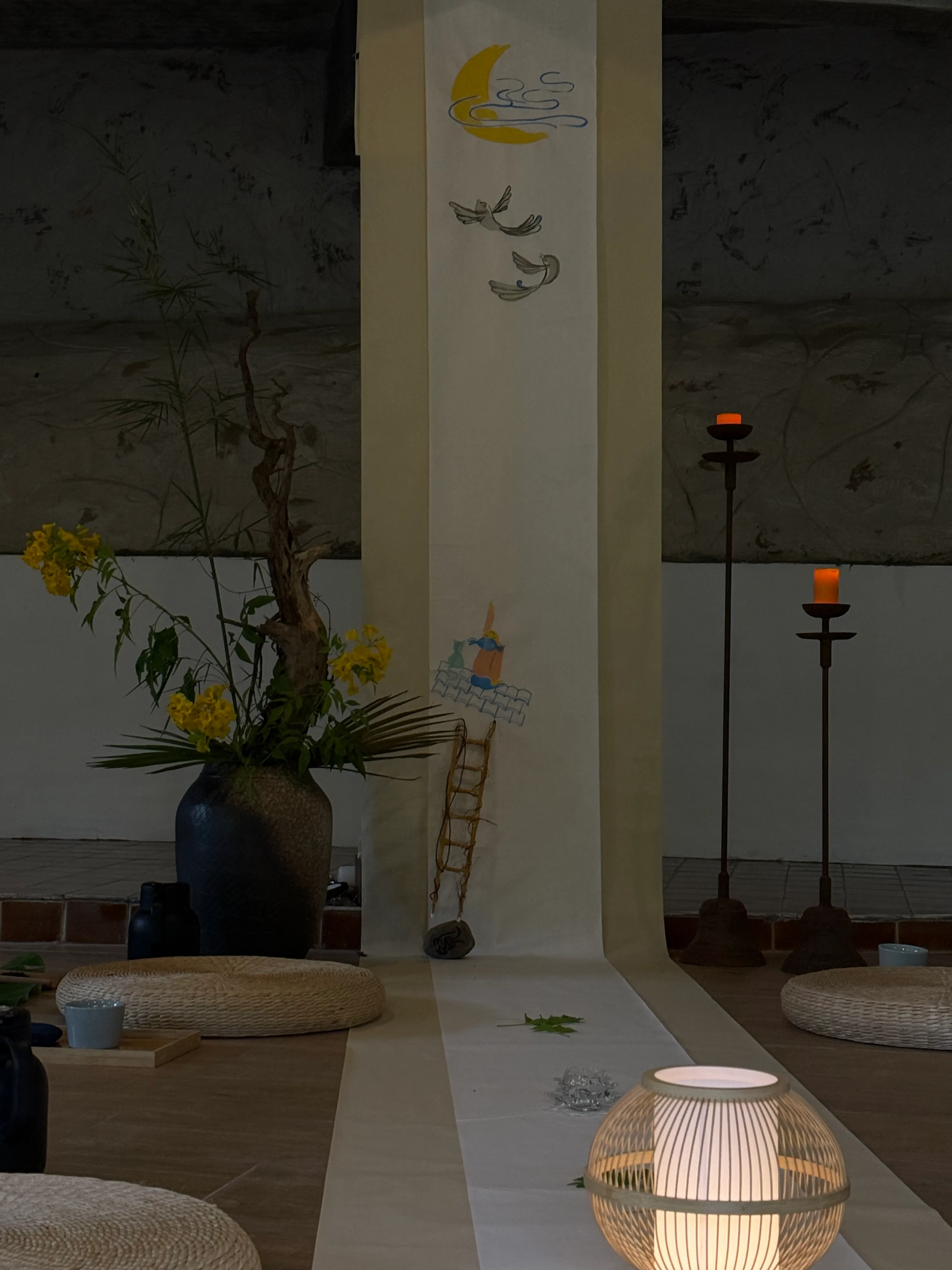 Meditation space with yellow flowers in dark vase, moon and feather illustrations on pillar, woven lanterns, and black candle holders