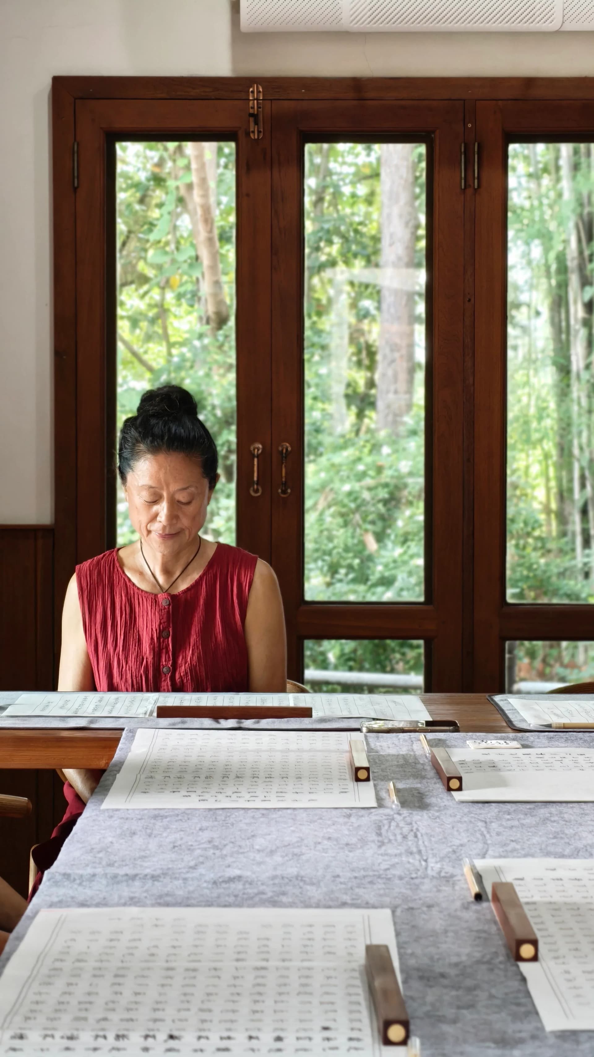 Woman in red sleeveless top sits at wooden table reviewing documents with forest views visible through large wooden-framed glass doors behind her
