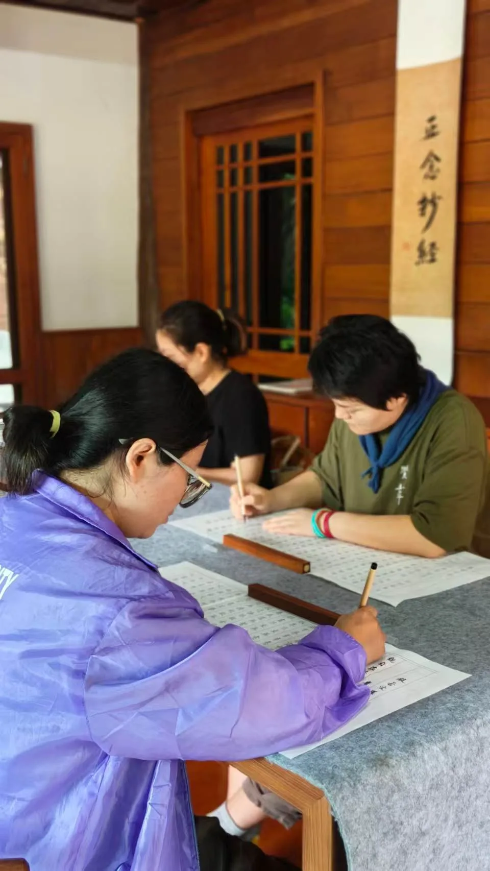 Multiple people practicing calligraphy with brushes at a wooden table indoors, with Japanese calligraphy artwork displayed on the wall