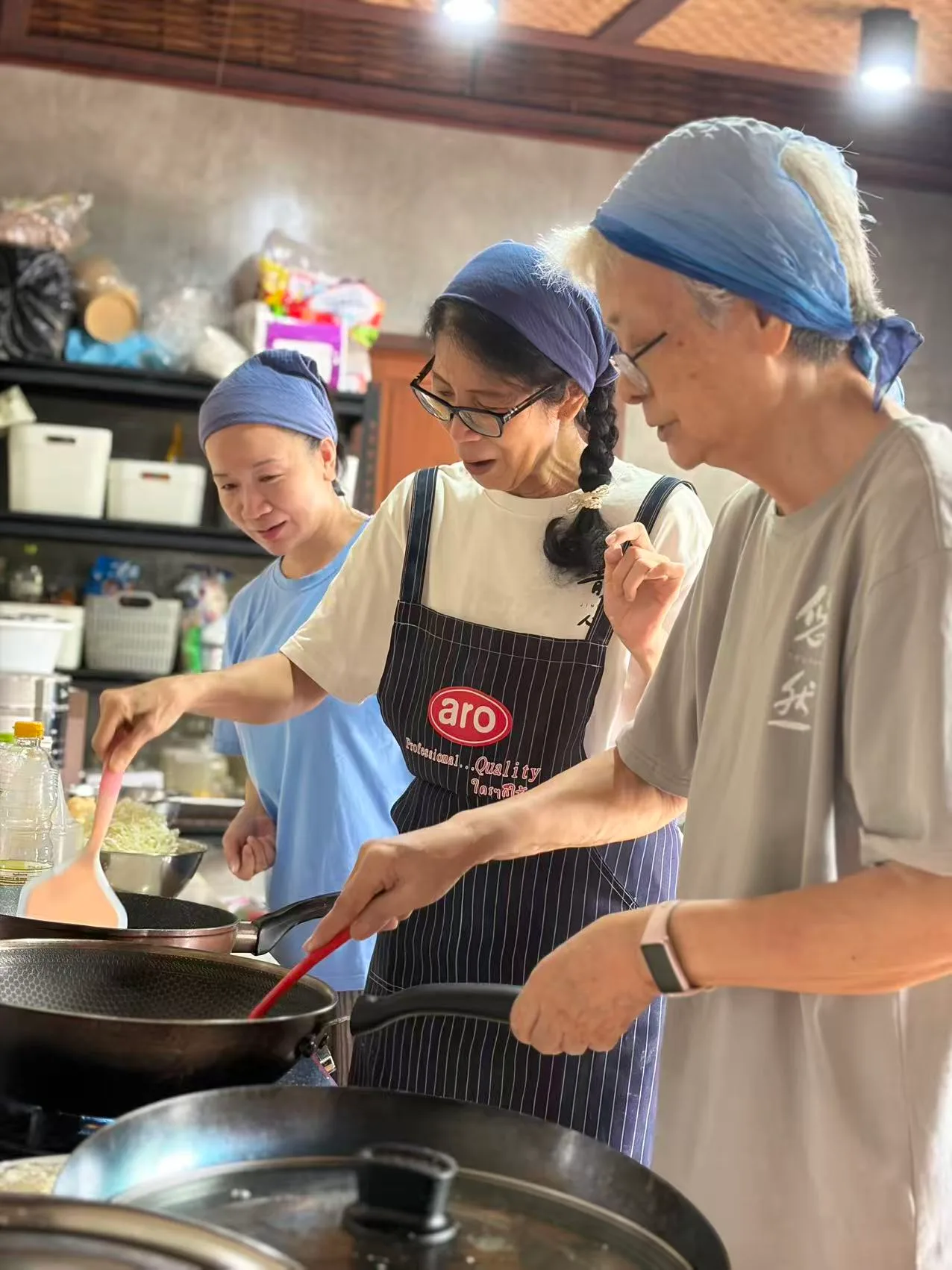 三人在厨房里一起烹饪，穿着蓝色头巾和围裙，在锅里翻炒食材