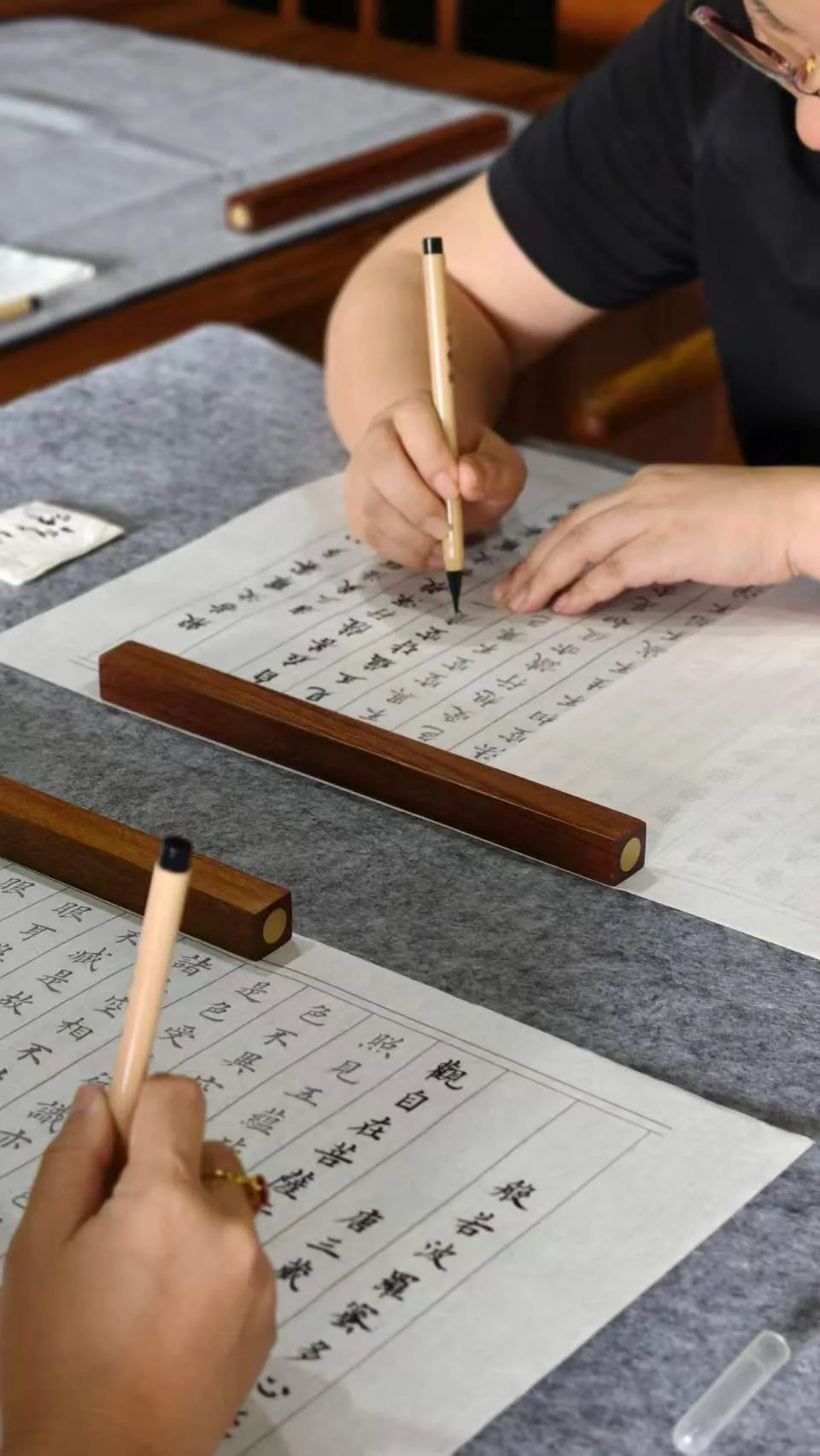 Two people practicing Chinese calligraphy with brushes on wooden brush rests at a table