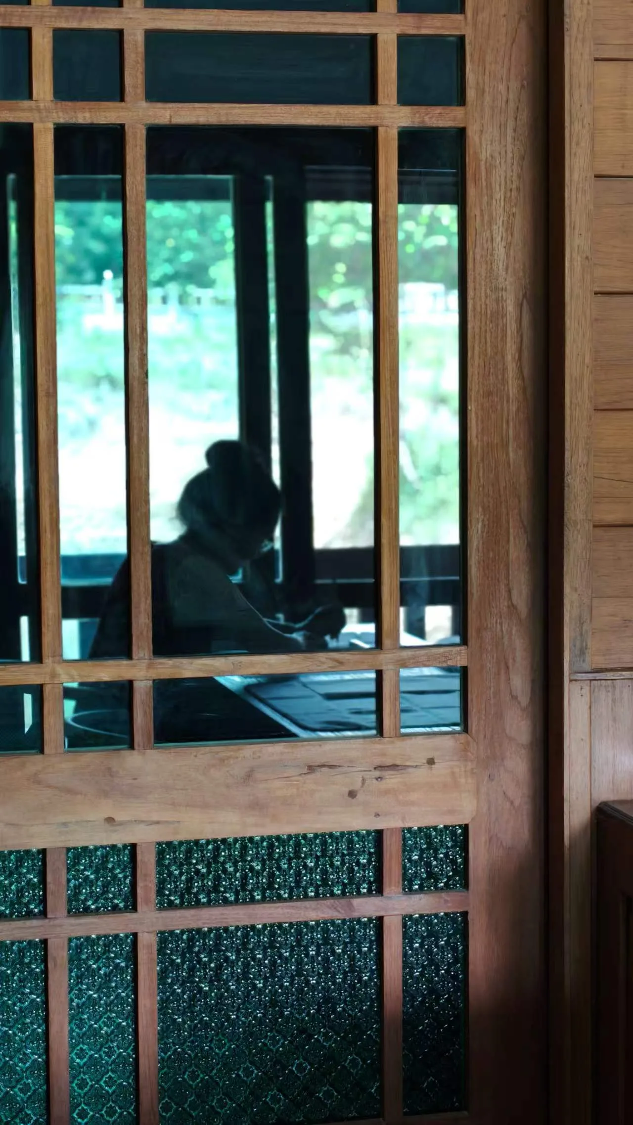 Person sitting indoors viewed through a wooden-framed window with green foliage visible outside