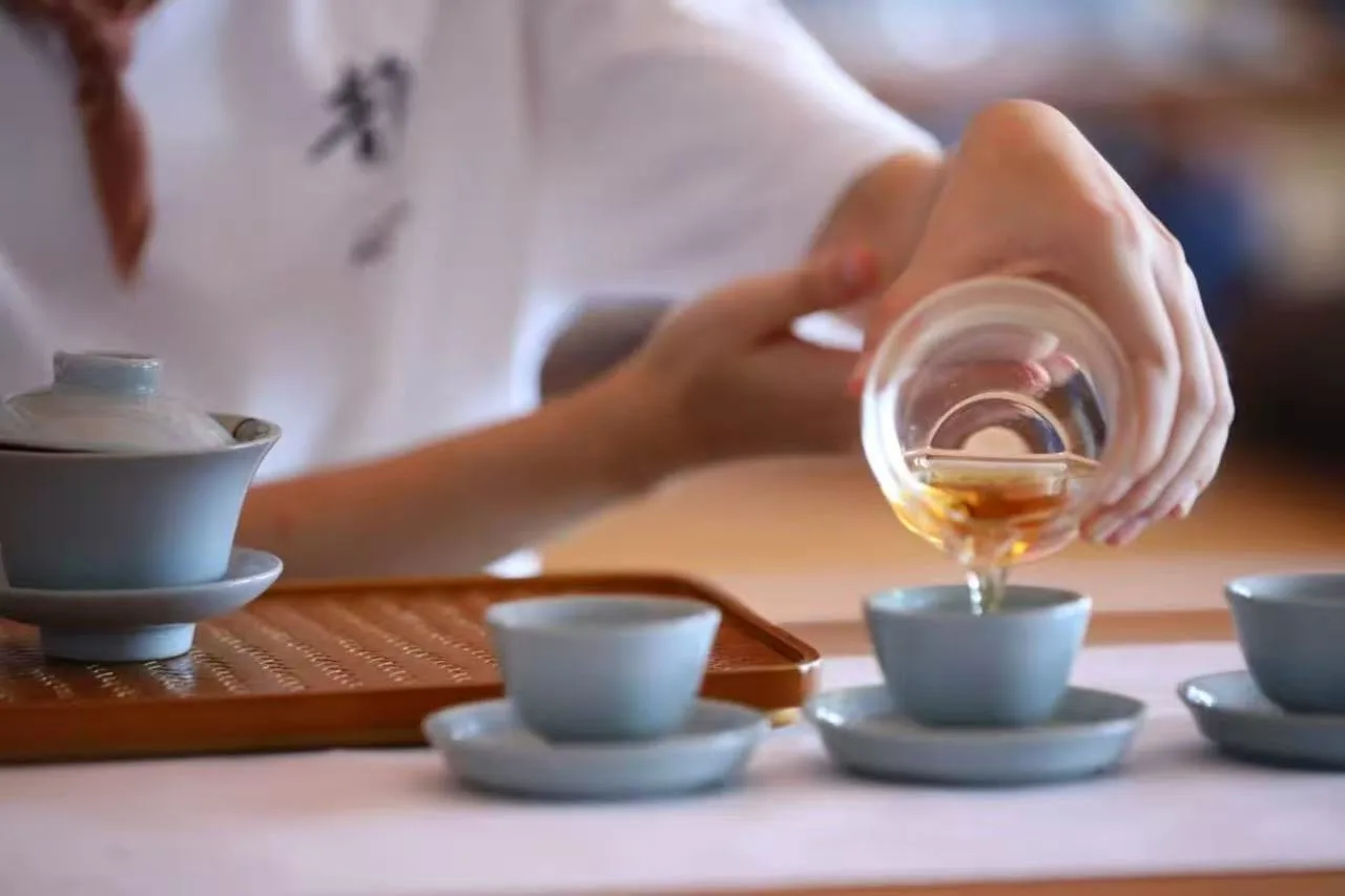 手持粉色茶壶向白瓷茶杯倒茶,茶具摆放在木制茶盘上