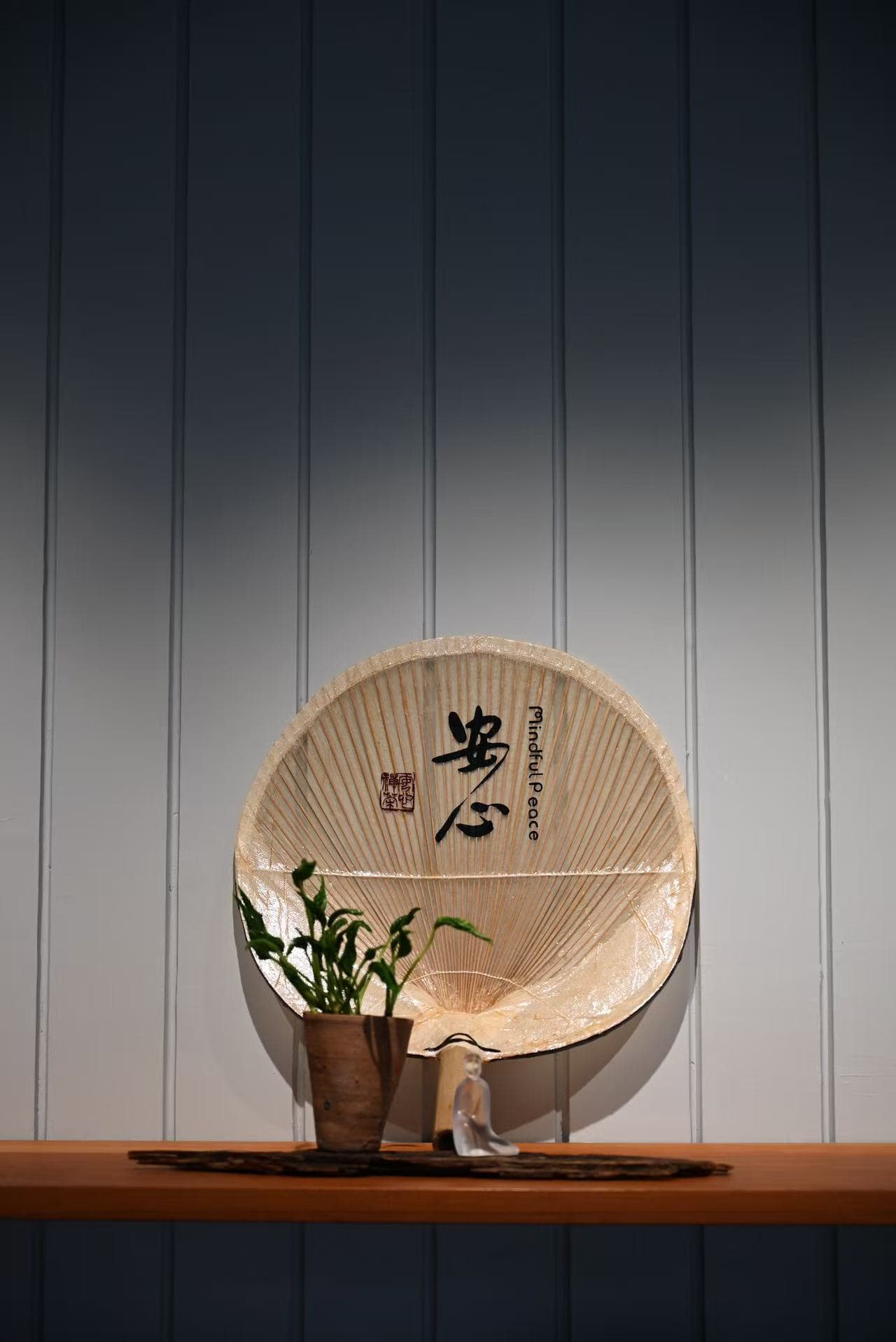 Circular wooden fan with Chinese characters displayed on wooden shelf alongside potted plant and small ceramic figurine against vertical striped wall