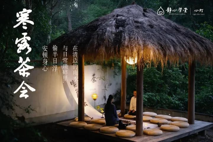 Person seated on cushion inside tea pavilion with wooden posts, lantern lighting, white walls with calligraphy, and surrounding greenery