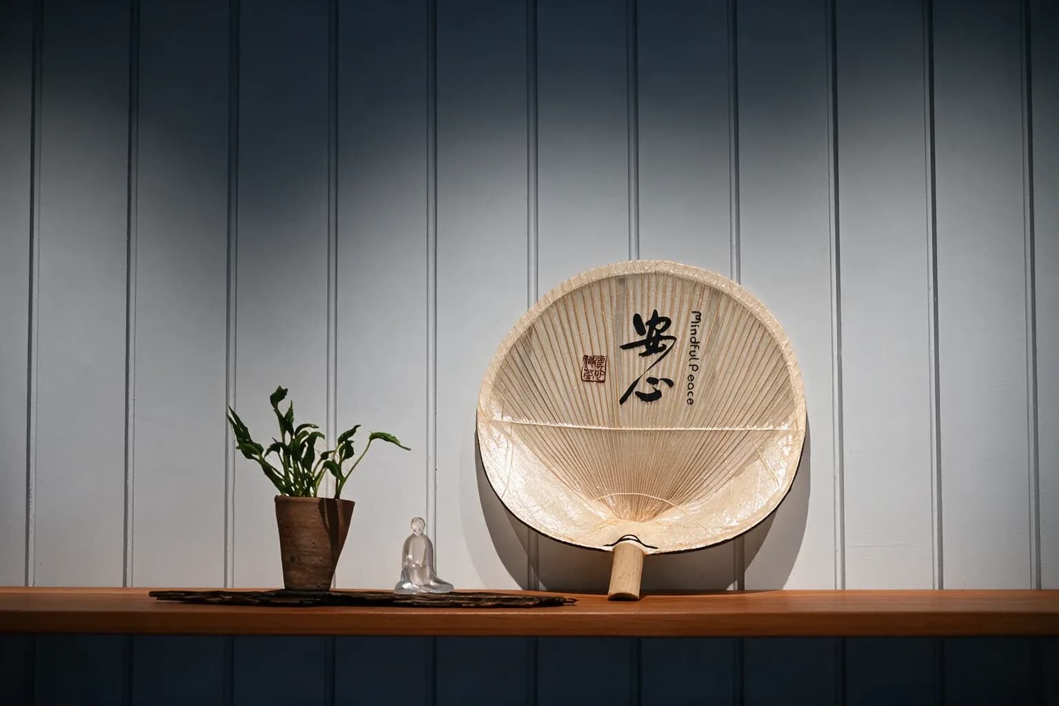 Circular wooden fan with Chinese characters displayed on wooden shelf alongside potted plant and small ceramic figurine against vertical striped wall