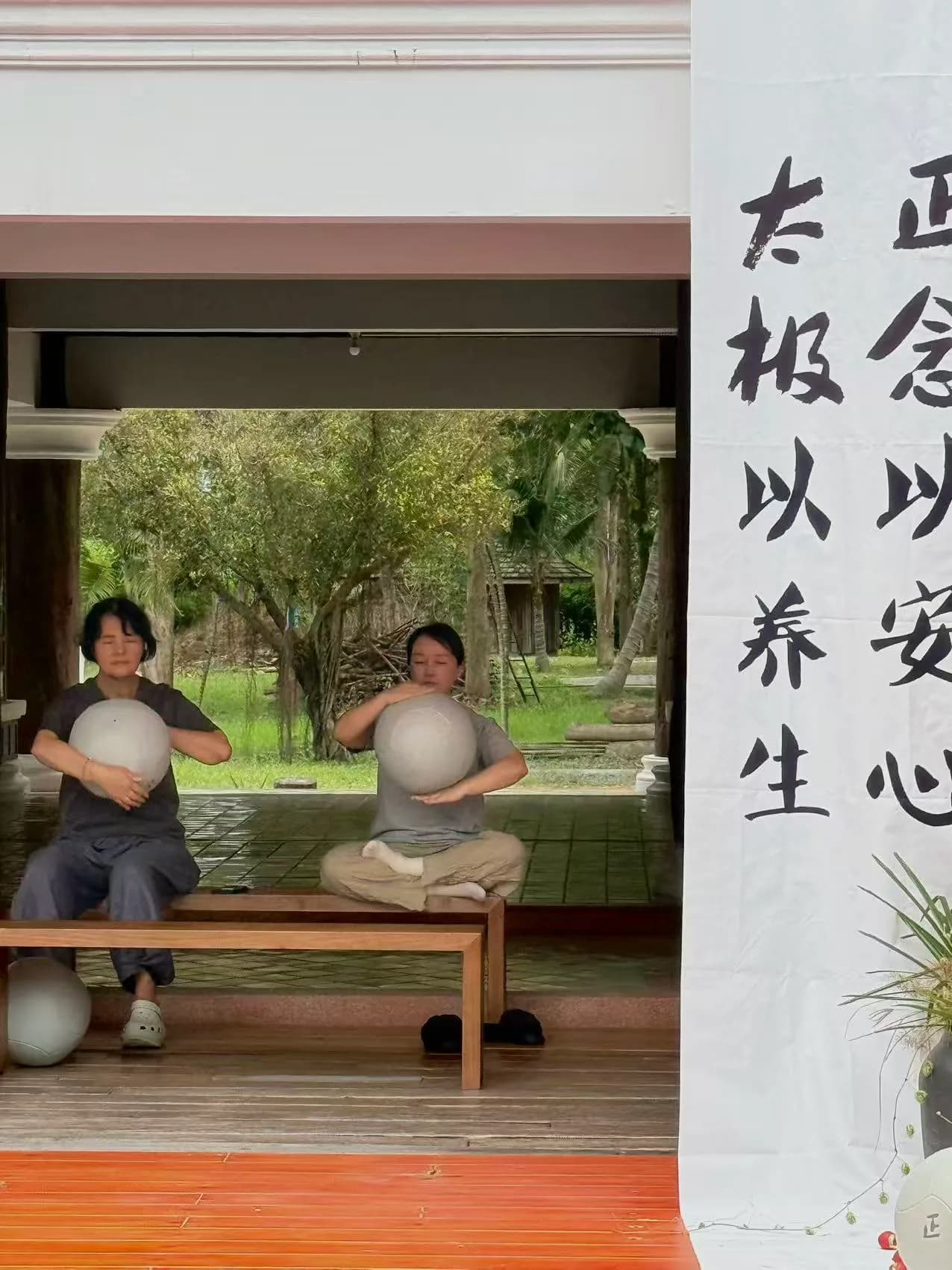 Two people sit on a wooden platform practicing with exercise balls, with lush greenery and traditional architecture visible behind them, and Chinese calligraphy on the wall to the